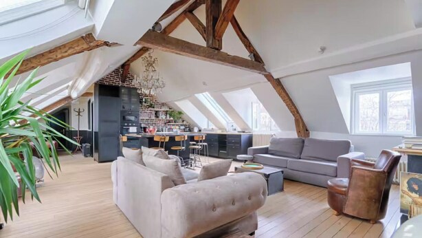 A spacious loft with a high ceiling, exposed wooden beams, a kitchen area, and a seating area with sofas and a coffee table.