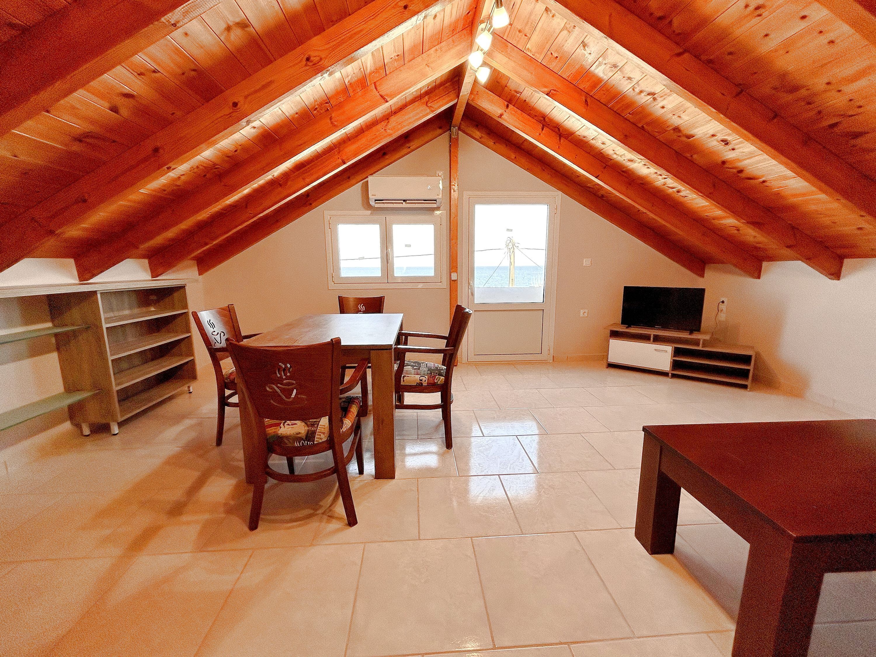 A spacious attic room with a wooden ceiling, a dining table with chairs, a TV stand, and a window with a view.