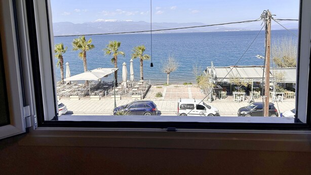 A view from a window overlooking a coastal area with palm trees, a parking lot, and a body of water. - Bella Vista Junior in Kamena Vourla (Kamena Vourla)