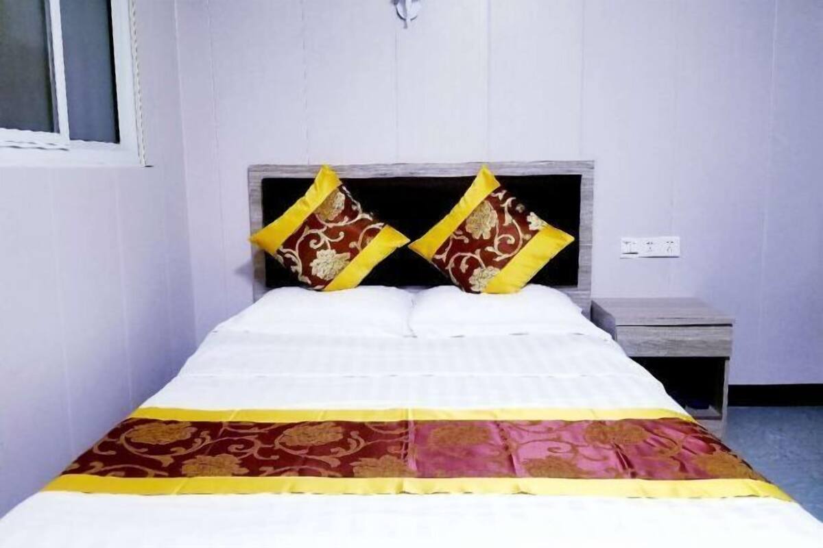 A bed with a white and patterned bedspread, two decorative pillows, a nightstand, and a wall socket.