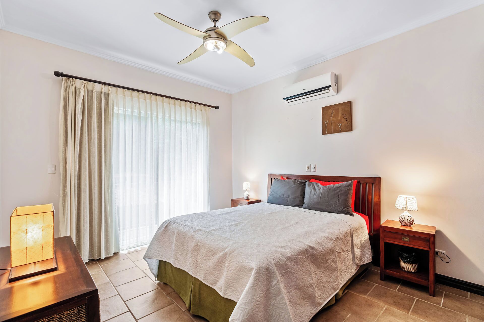 Quarto de hotel com cama, mesas de cabeceira, ventoinha no teto, ar condicionado e janela com cortinas.