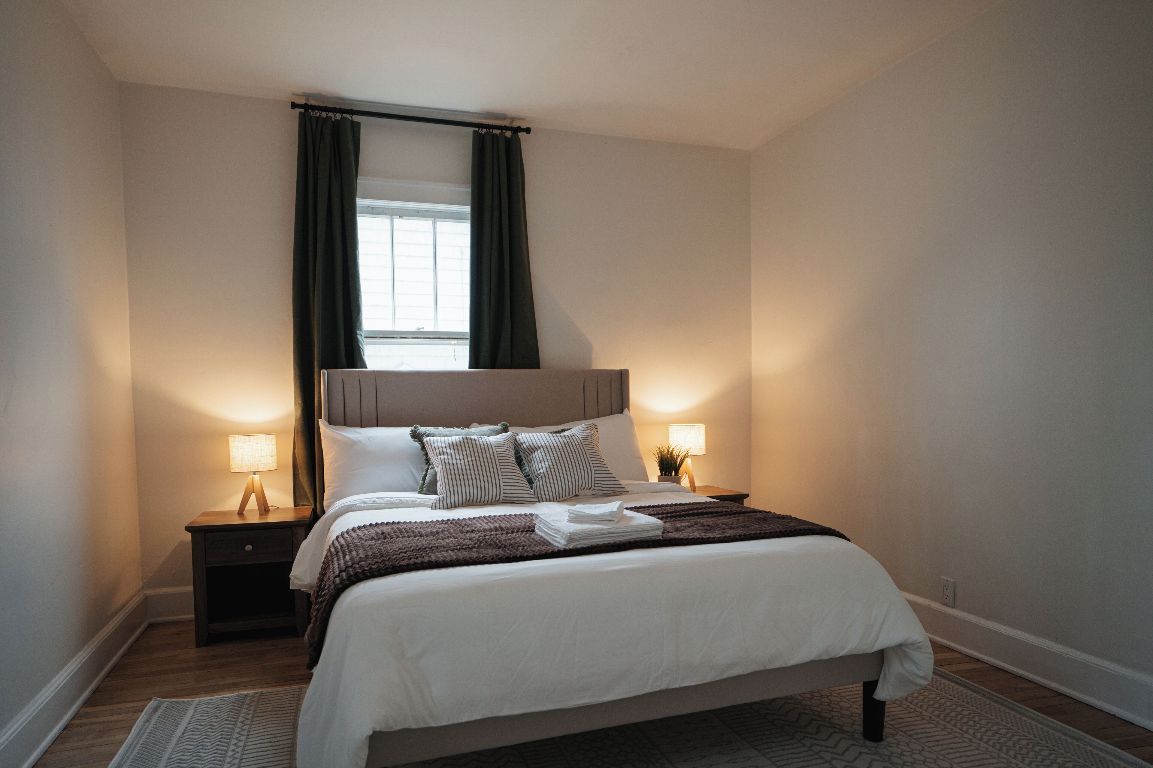 A neatly arranged bedroom with a bed, bedside lamps, a window with curtains, and a nightstand.
