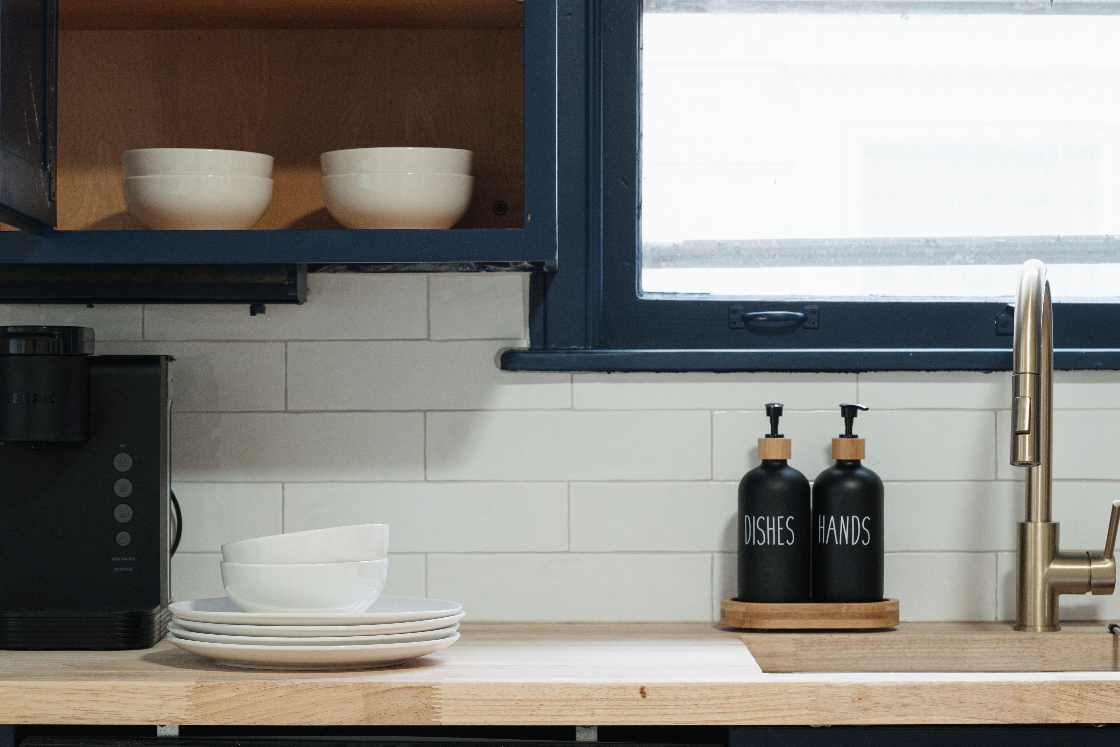 A kitchen with white tiles, a black coffee machine, white bowls, and black dish soap bottles.