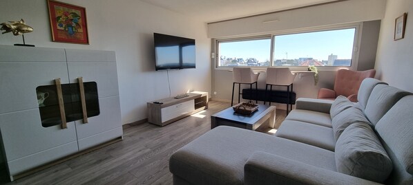 Una sala de estar moderna con un sofá, una mesa de centro, un televisor montado en la pared y un amplio ventanal con vista a la ciudad.