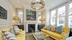 A bright living room with yellow sofas, a fireplace, and large windows offering a view of the outside. - Elegant 4 Bedroom House - Clapham (London)