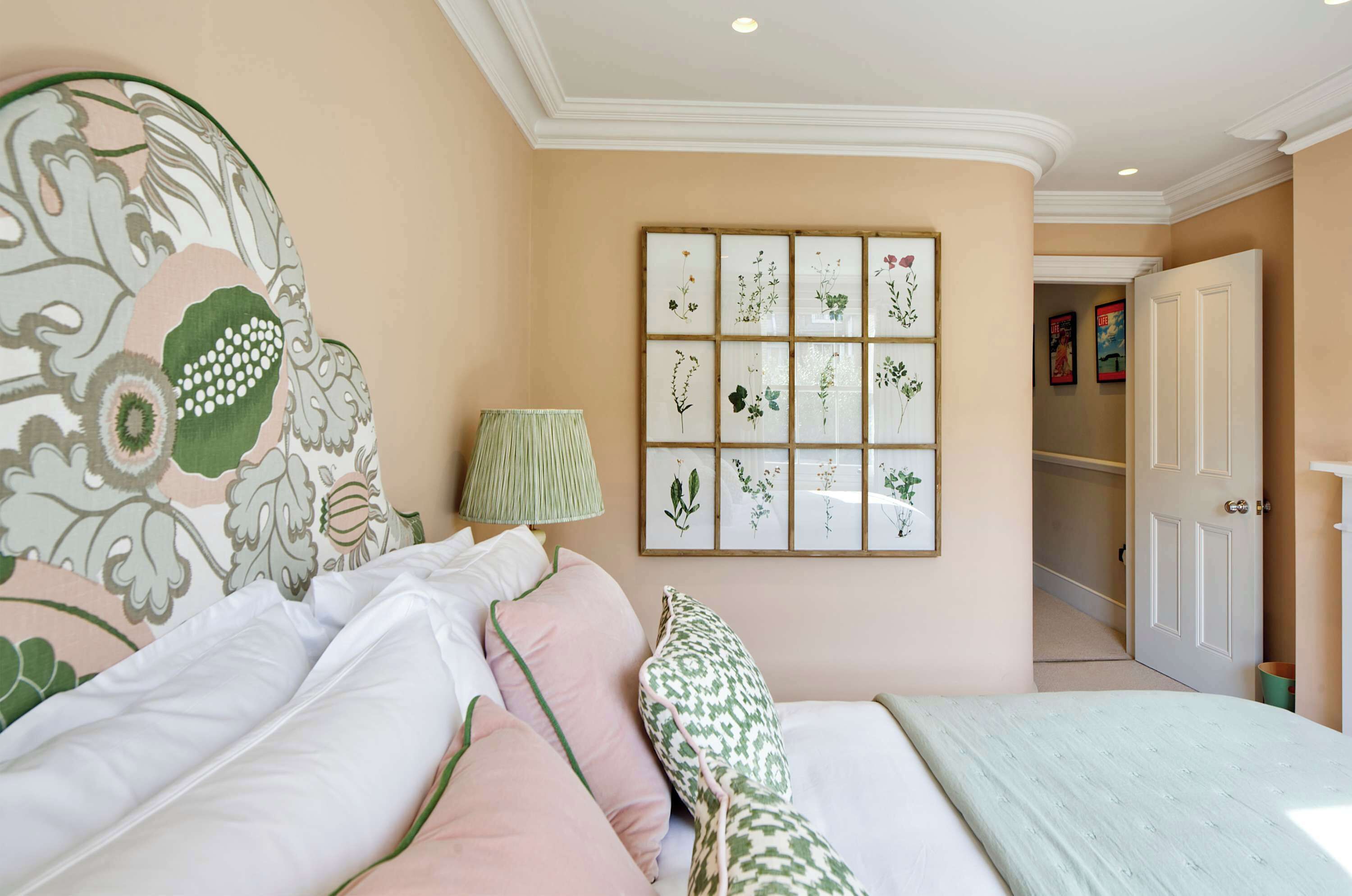 A bedroom with a large bed, a bedside table with a lamp, and a wall with framed botanical prints.