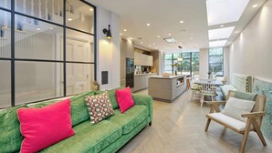 A modern living room with a green sofa, wooden chairs, and a kitchen in the background. - Elegant 4 Bedroom House - Clapham (London)