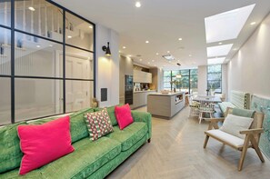 A modern living room with a green sofa, wooden chairs, and a kitchen in the background. - Elegant 4 Bedroom House - Clapham (London)