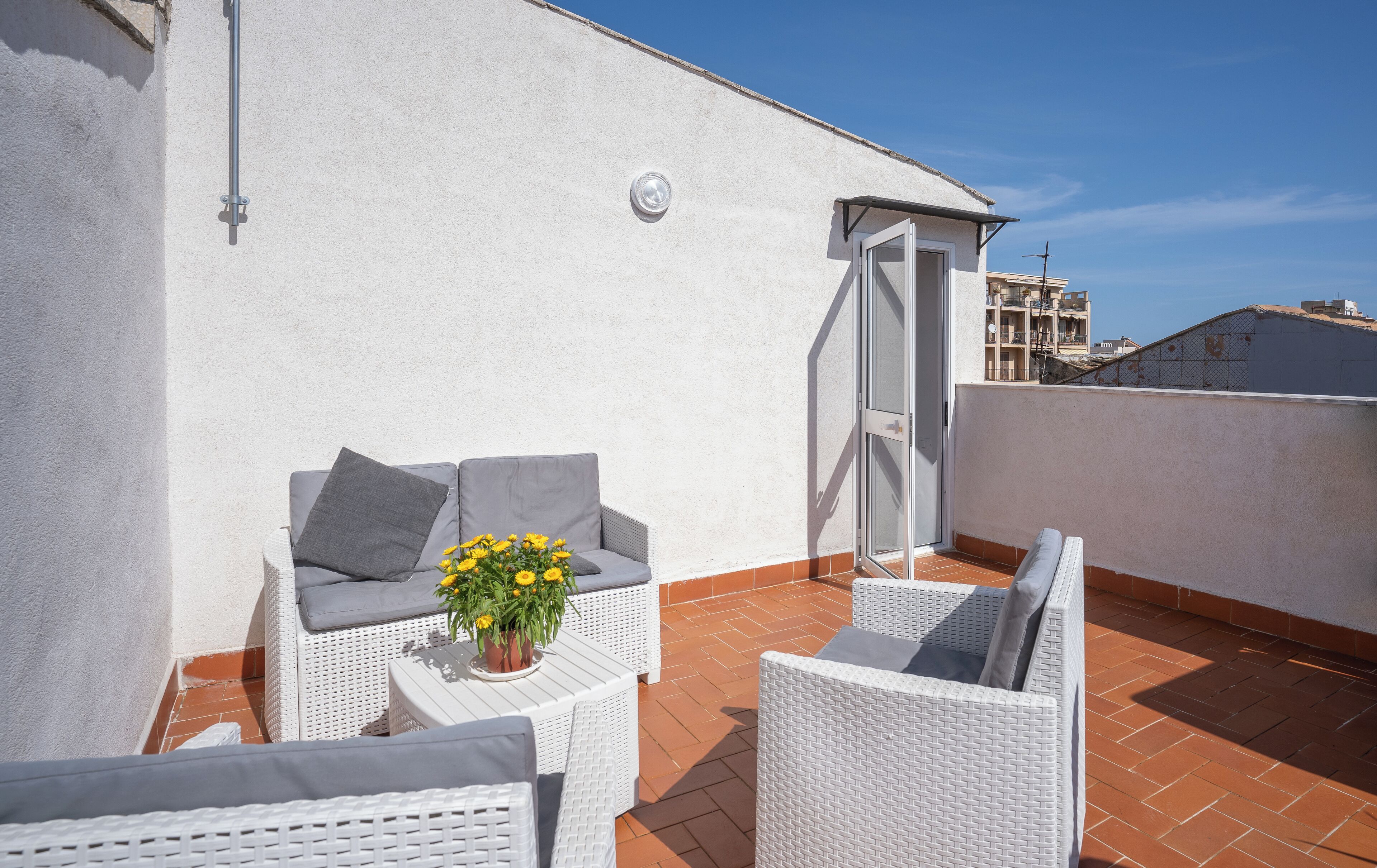 A rooftop terrace with white wicker furniture, a potted plant, and a door leading to an adjacent building.