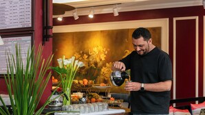 Una persona sirve una bebida en un vaso en la zona de recepción de un hotel, con un panel de menú, un arreglo floral y un área de estar al fondo.