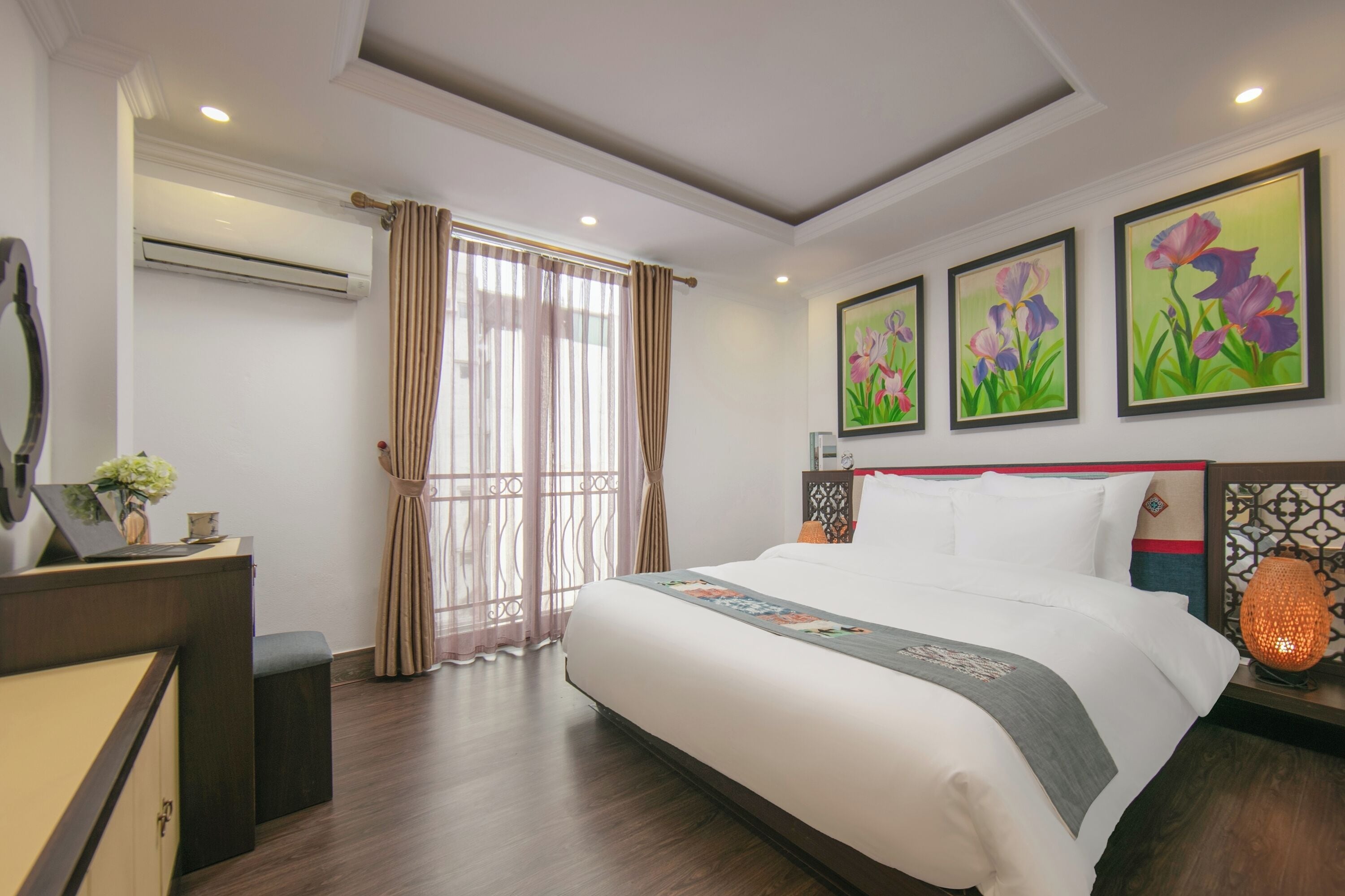 A hotel room with a large bed, a desk with a mirror, and three framed floral paintings on the wall.