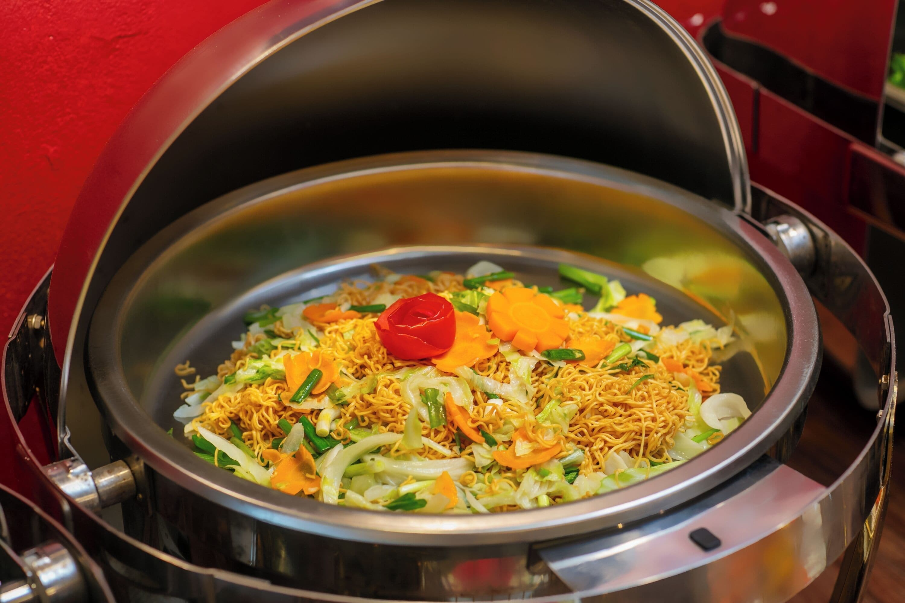 Una olla de acero inoxidable con tapa, conteniendo un plato de fideos, verduras y un adorno rojo.