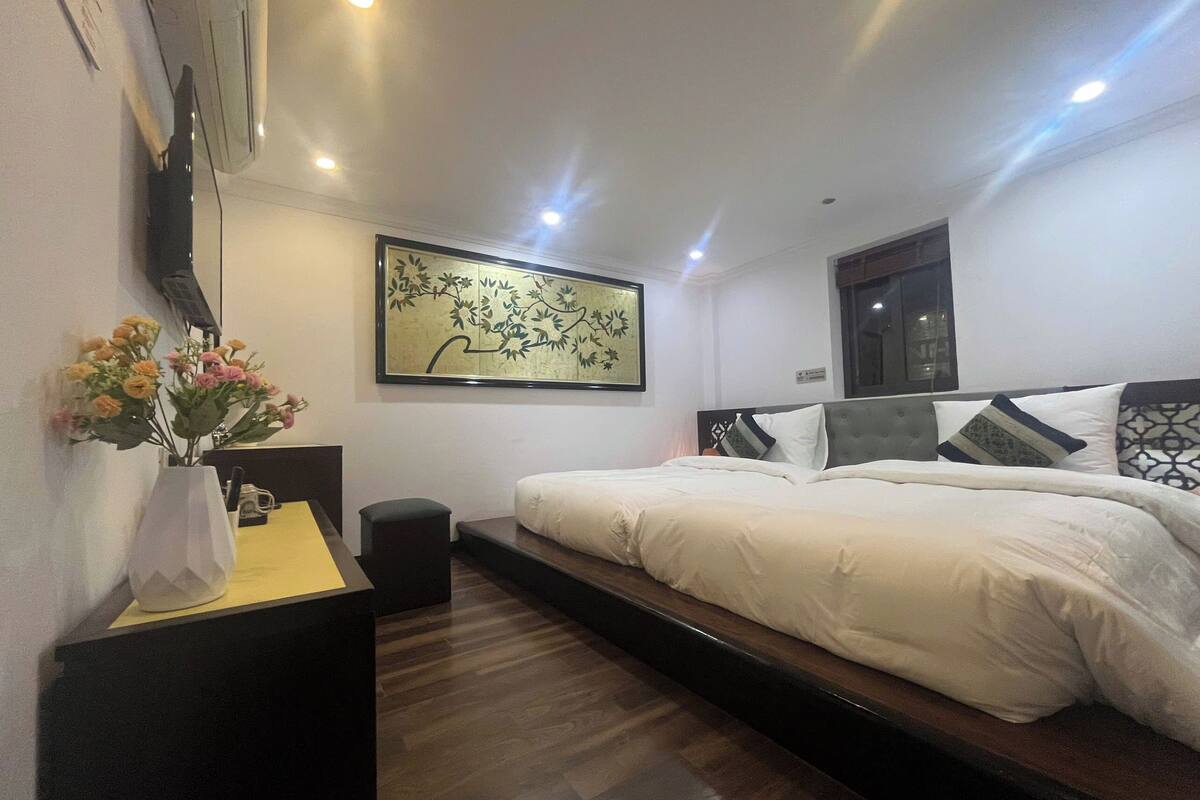 A modern bedroom with a large bed, a small desk, a vase with flowers, and a wall-mounted artwork.