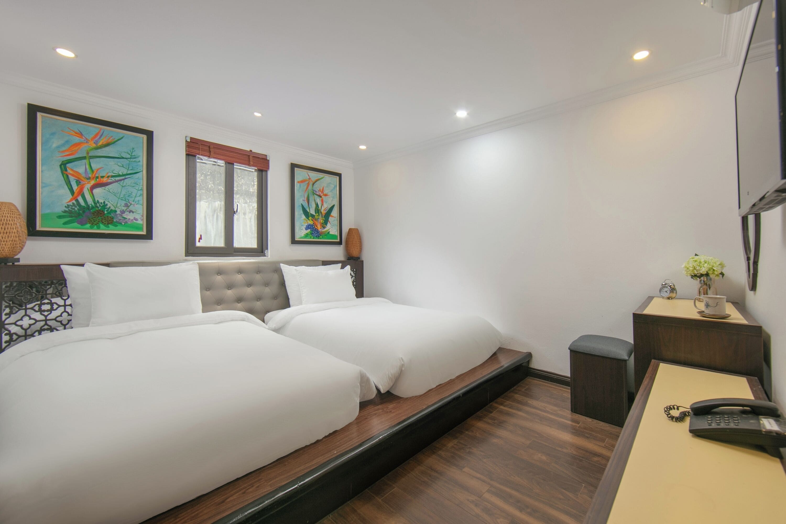 A hotel room with two beds, a desk with a telephone, and artwork on the walls.