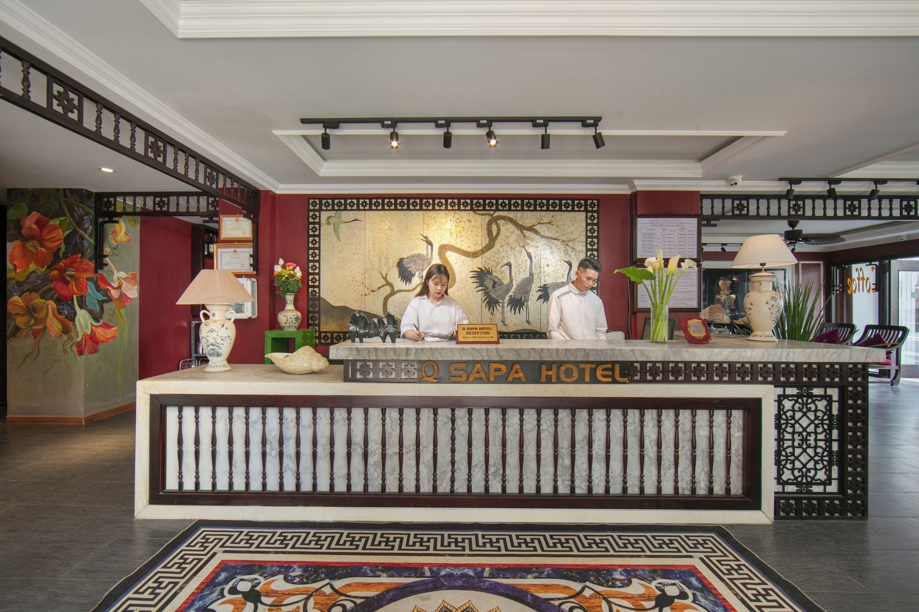 A reception desk at Q Sapa Hotel with a decorative mural, a nameplate, and a traditional lamp.
