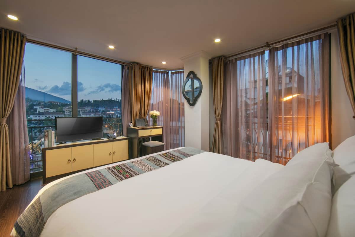 A hotel room with a large bed, a TV on a cabinet, a desk with a chair, and a view of a cityscape through large windows.