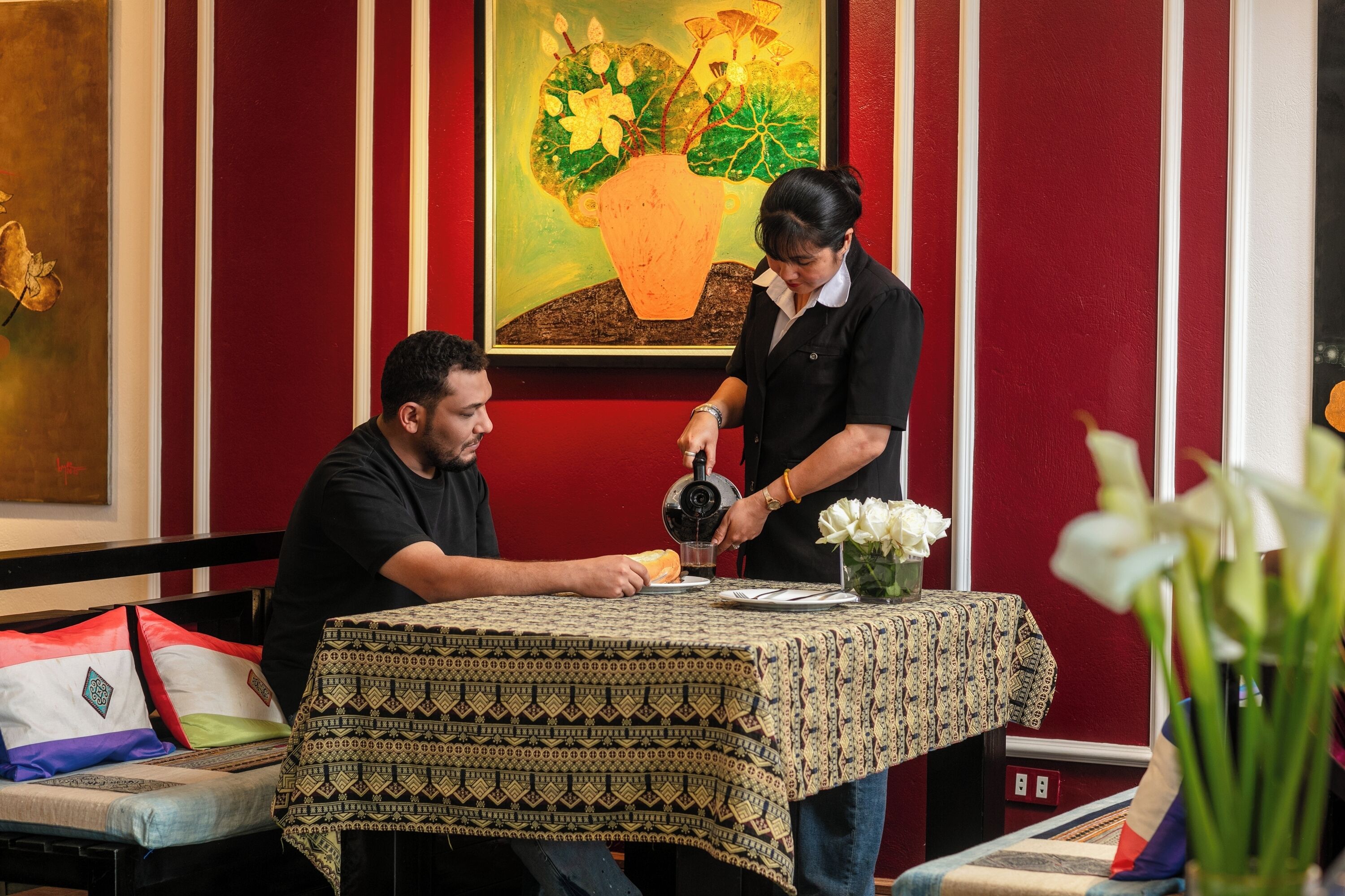 A man and a woman at a table with a patterned tablecloth, a vase with white flowers, and a painting of a plant on the wall.