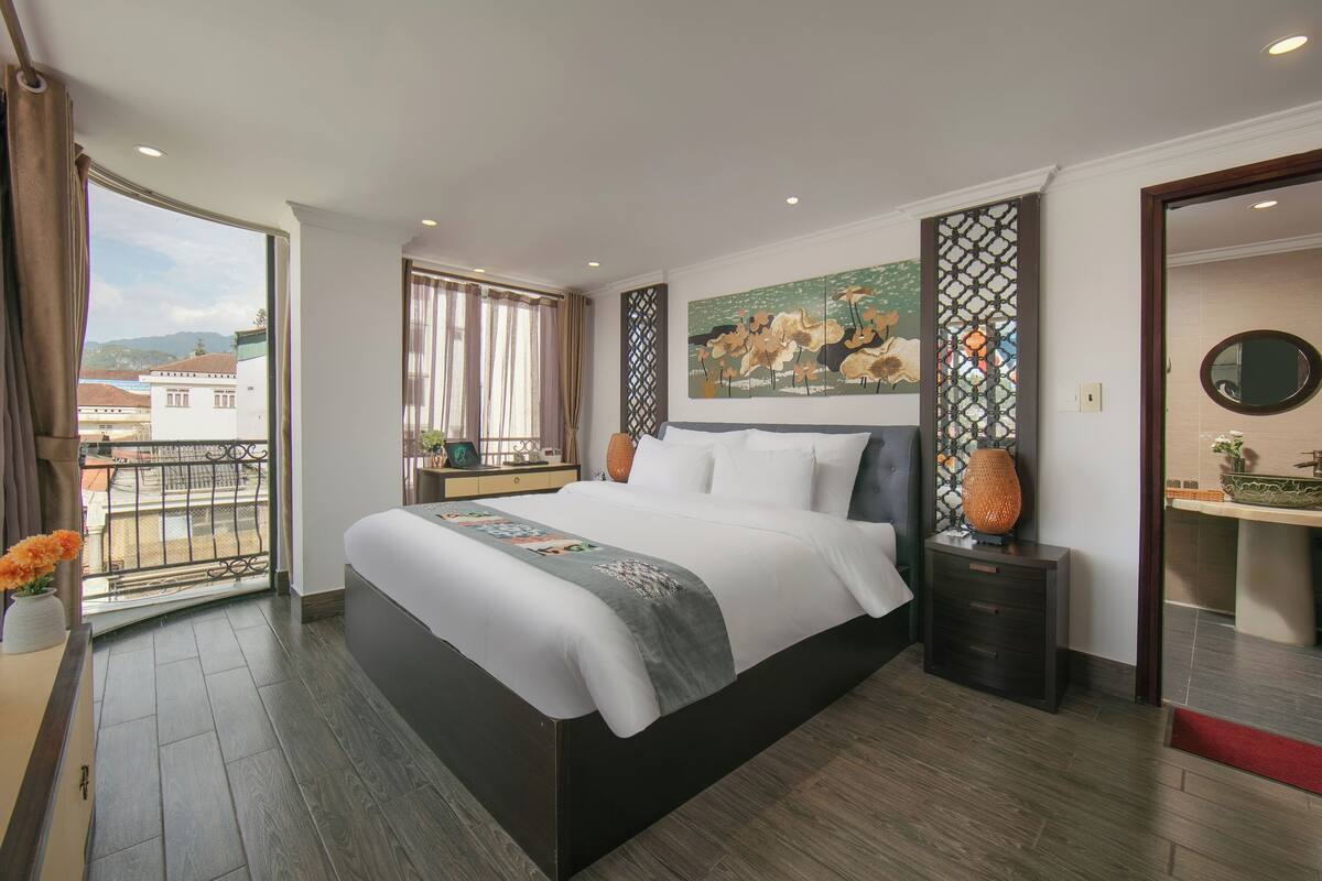 A modern hotel room with a large bed, a balcony with a view, a painting on the wall, and a bathroom with a round mirror.