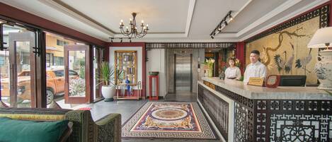 A hotel lobby with a reception desk, a chandelier, a patterned rug, and a view of the street outside.