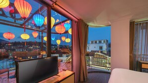 Habitación de hotel con un ventanal grande que da a un paisaje urbano, un televisor de pantalla plana y un balcón con mobiliario exterior.