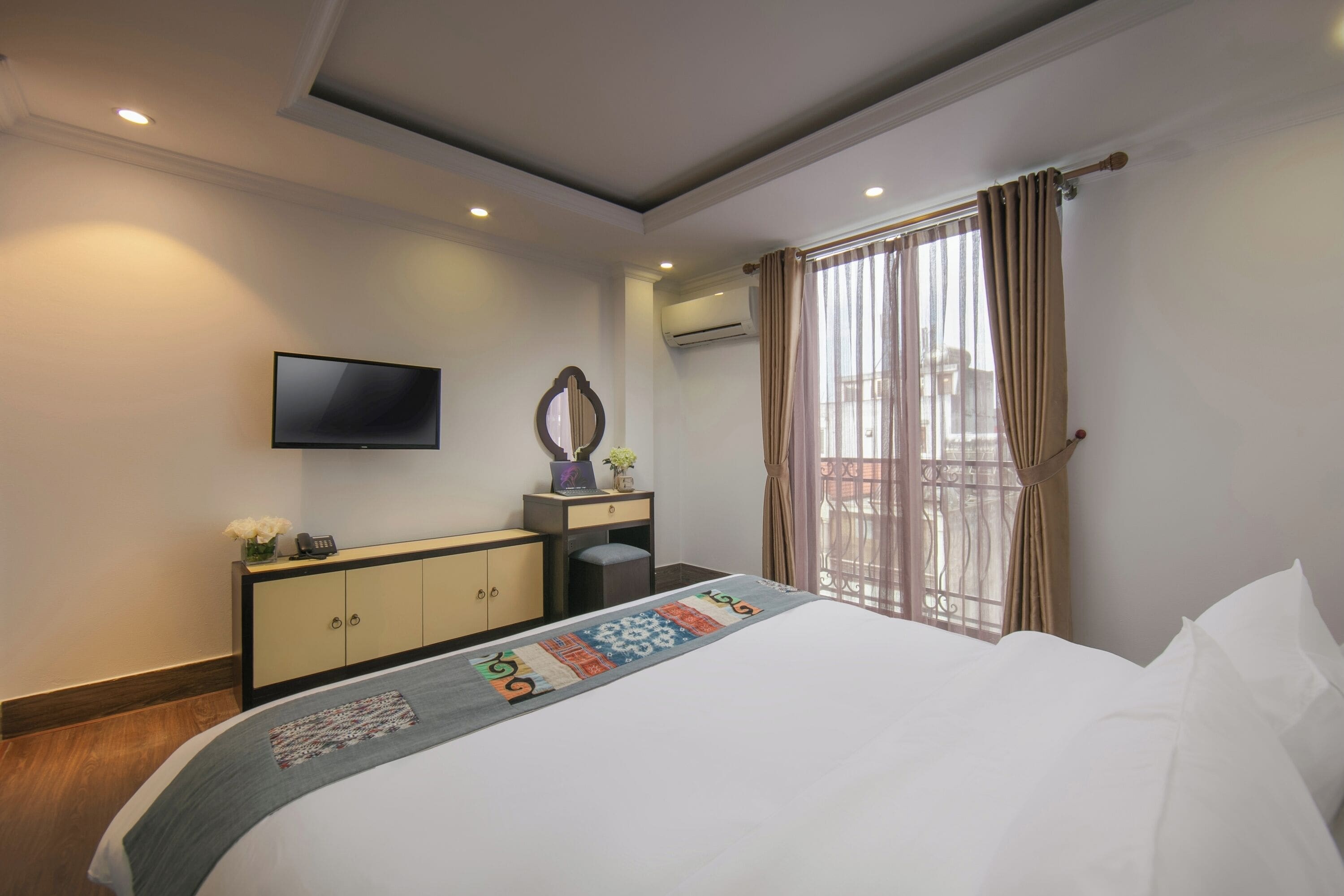 Habitación de hotel con cama, televisor montado en la pared, escritorio con espejo y ventana con cortinas.