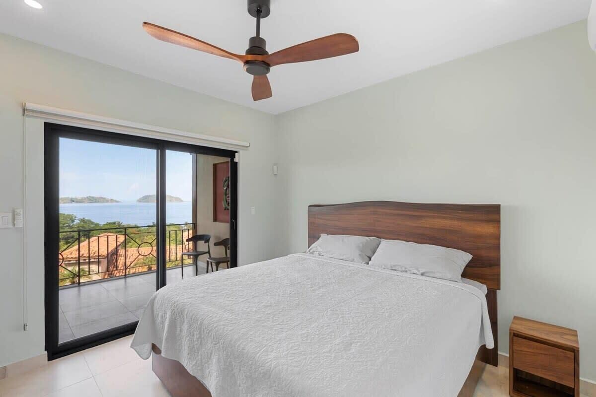 A bedroom with a large bed, a ceiling fan, a nightstand, and a view of the sea through a sliding door.