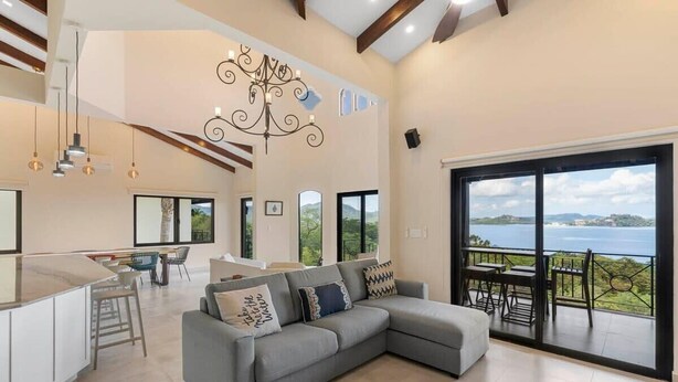 A spacious living area with a grey sectional sofa, a kitchen with a bar, and a balcony with a view of the sea. - Stylish Villa with Brand-New Furniture, Panoramic Ocean Views, and Infinity Swimming Pool & Hot Tub (Potrero)