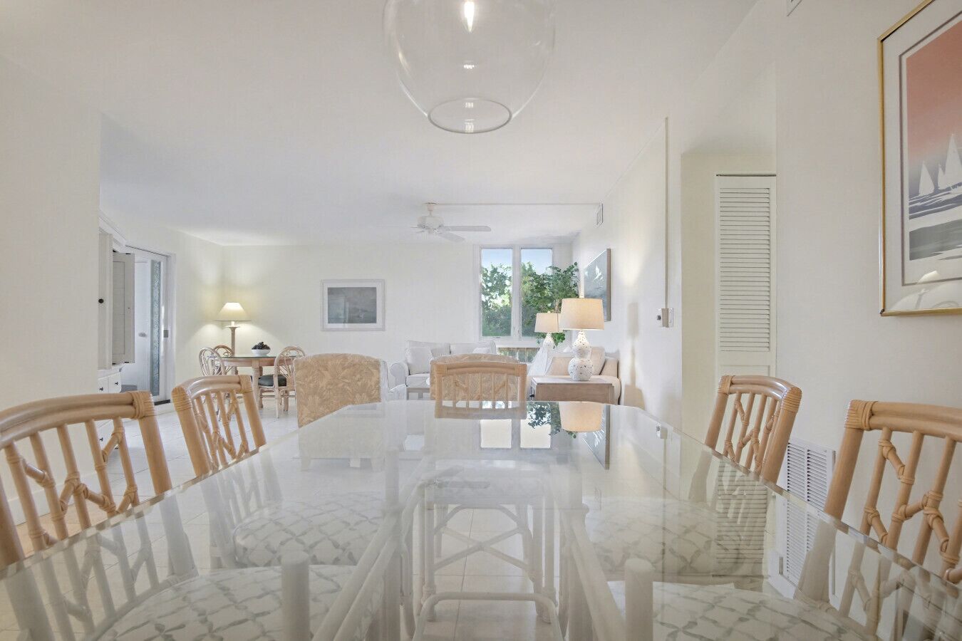 Uma ampla área de jantar com uma grande mesa de vidro rodeada por cadeiras de madeira, um sofá branco e uma pequena mesa lateral com uma luminária.