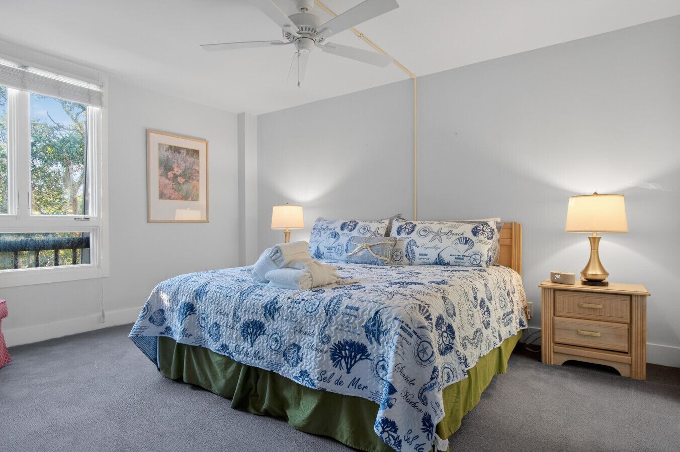 Un dormitorio con cama, dos mesitas de noche, ventilador de techo y ventana con vista a árboles.