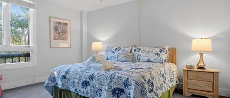 Un dormitorio con cama, dos mesitas de noche, ventilador de techo y ventana con vista a árboles.