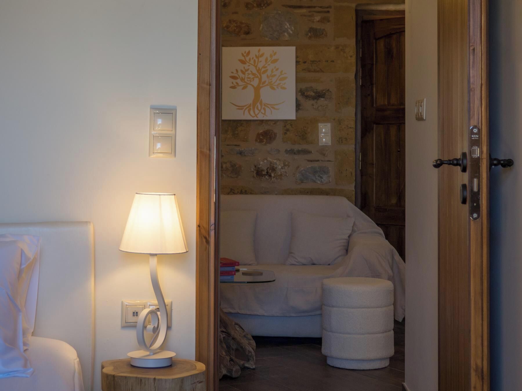 Una habitación acogedora con un sofá blanco, una mesita redonda de madera con una lámpara, una pared de piedra con un mural de un árbol y una puerta de madera.