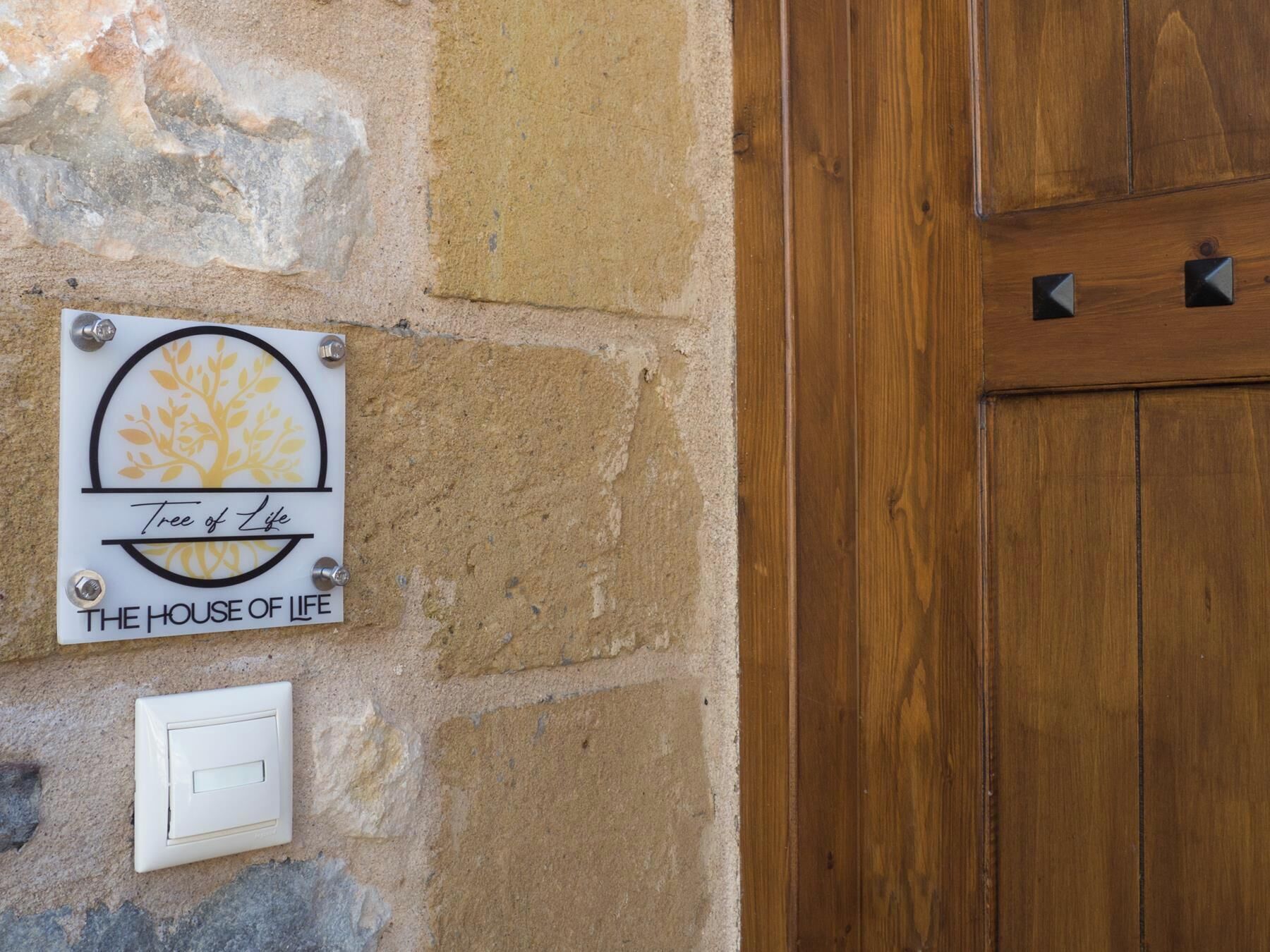 A sign with a tree and sun design, labeled 'Tree of Life' and 'The House of Life' on a stone wall.