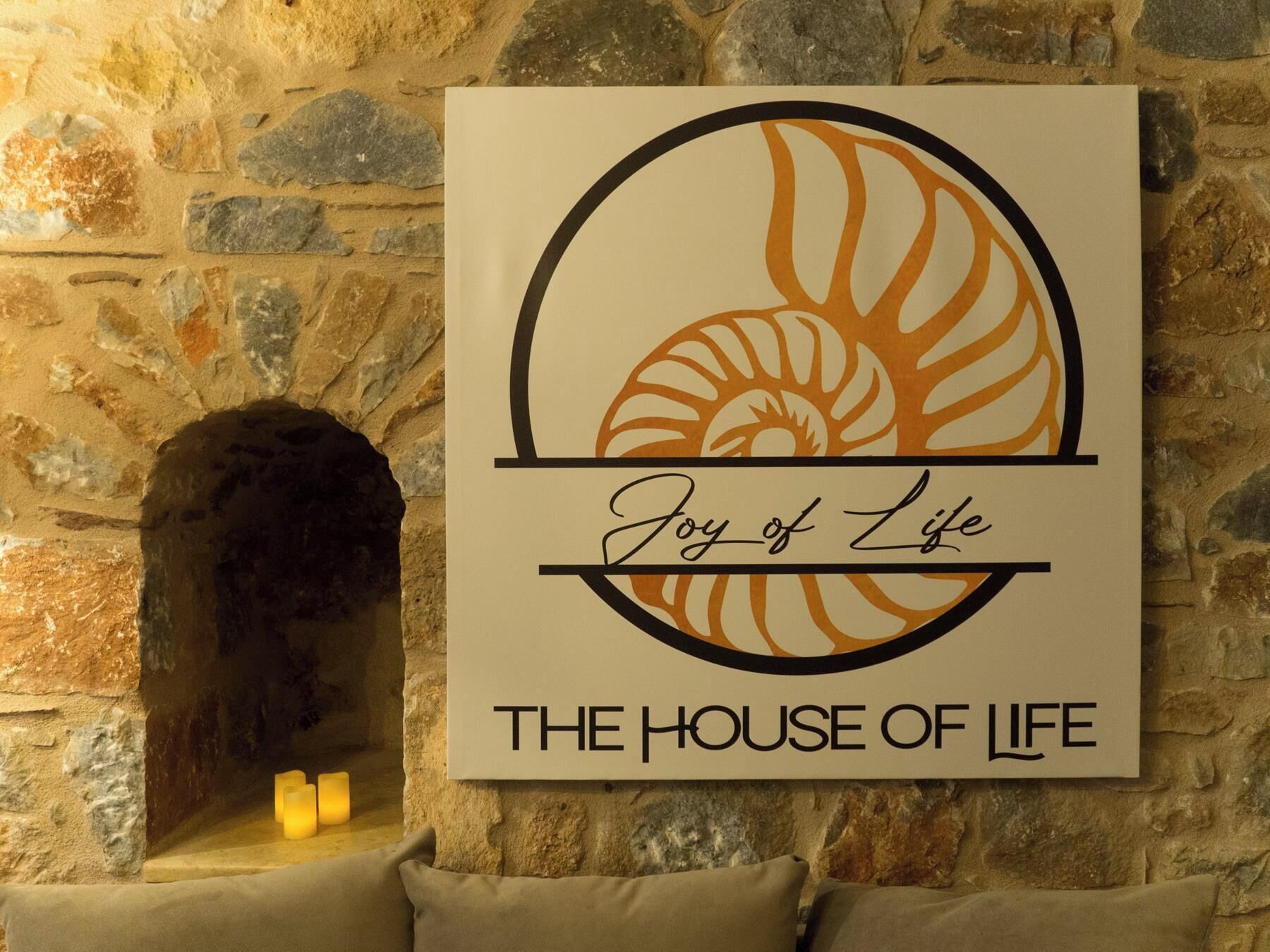 A wall-mounted sign with a circular design and text stating 'Joy of Life THE HOUSE OF LIFE' against a stone wall backdrop.
