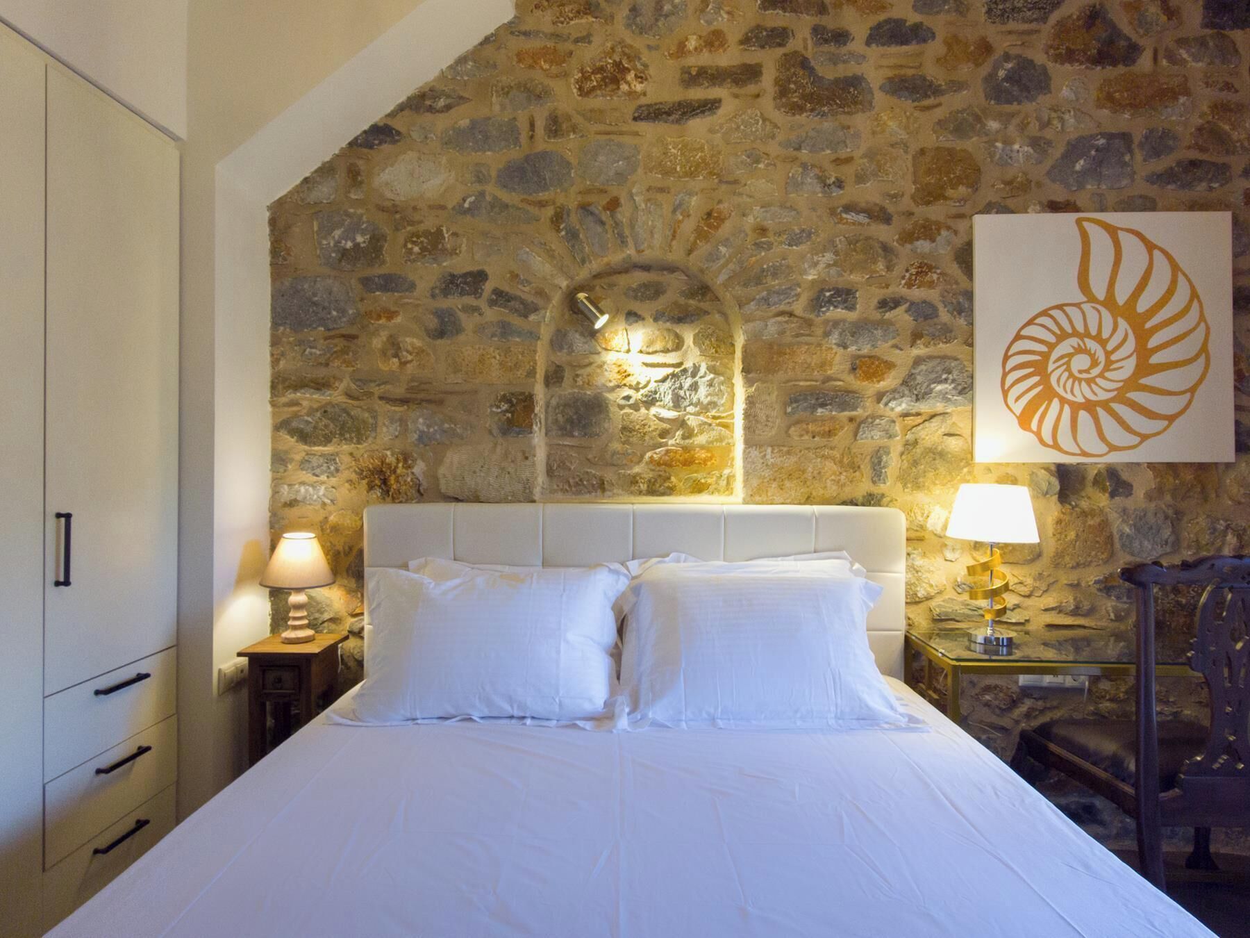 A bedroom with a stone wall, a bed with white linens, a nightstand with a lamp, and a framed artwork of a shell.