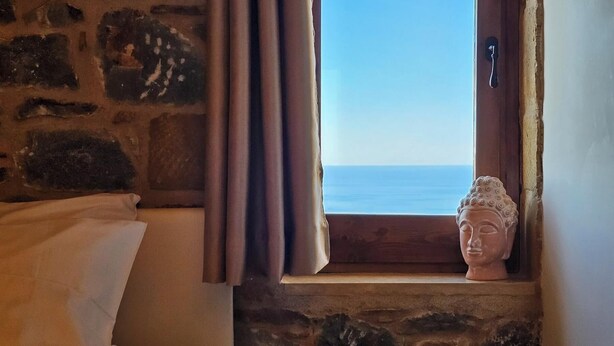 A stone wall, a window with a view of the sea, and a Buddha statue on a ledge.