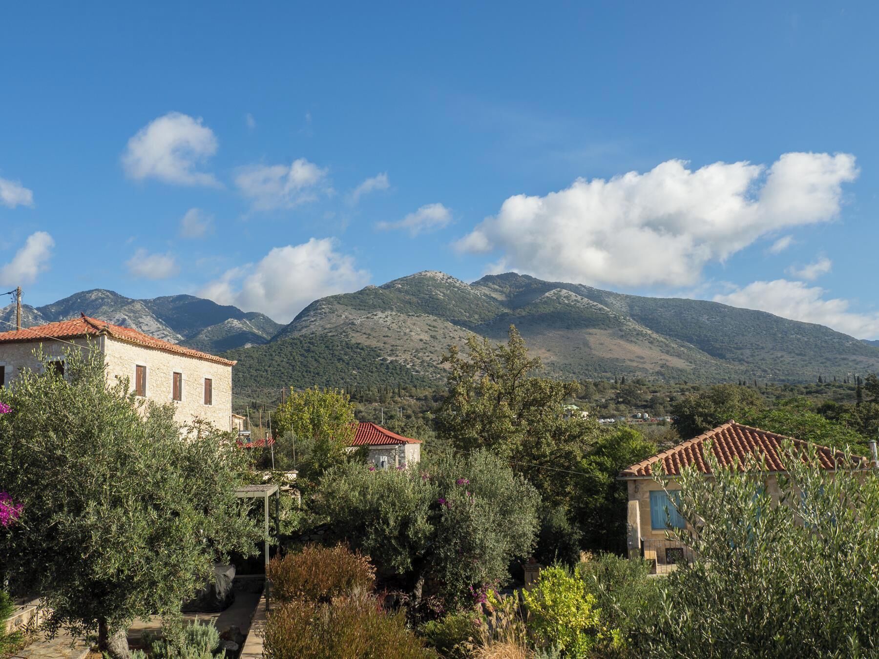 A traditional stone building with a red-tiled roof, surrounded by lush greenery and olive trees, with mountains in the background.
