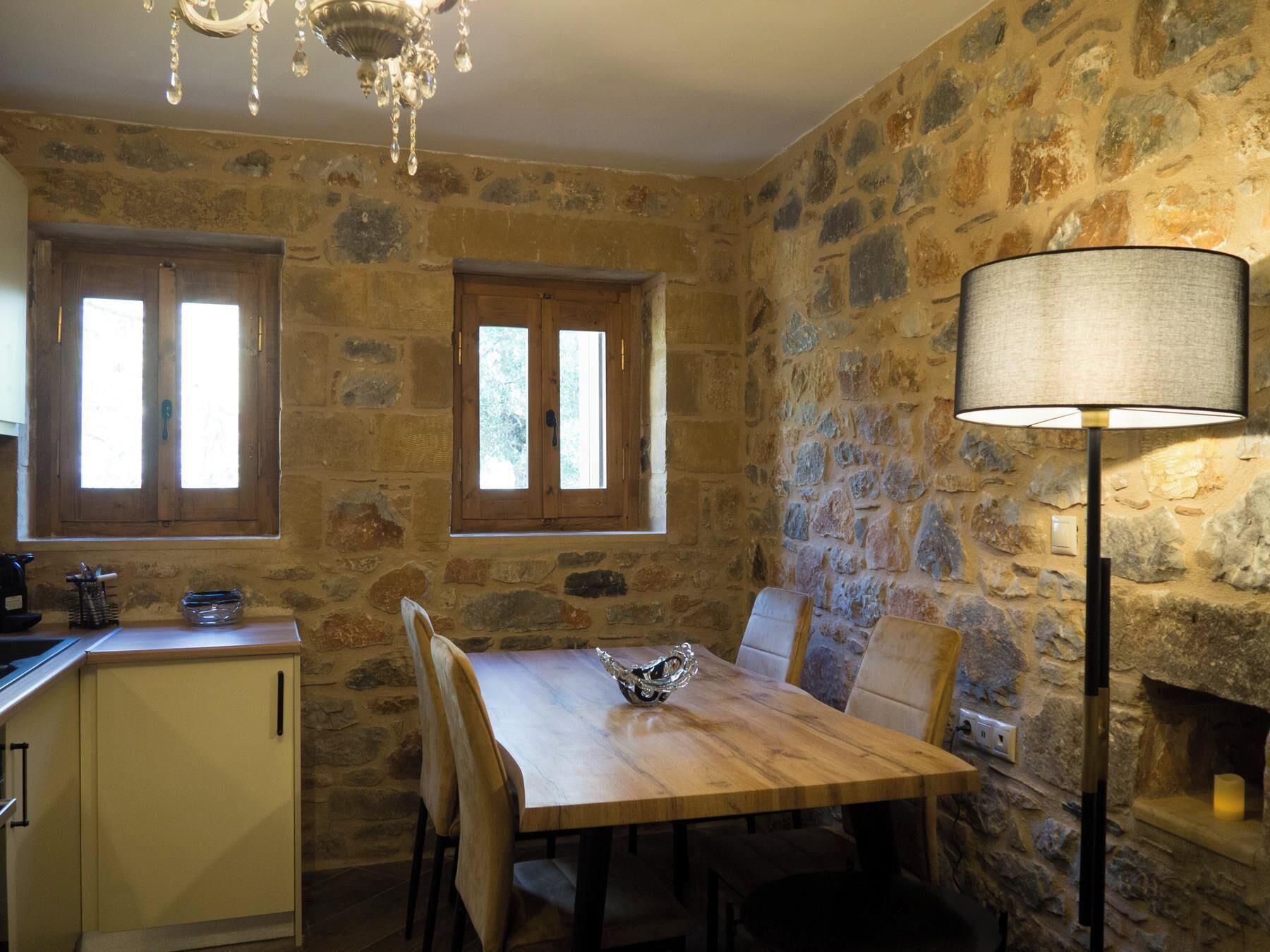 A rustic stone-walled room with a wooden dining table, two windows, a chandelier, and a floor lamp.
