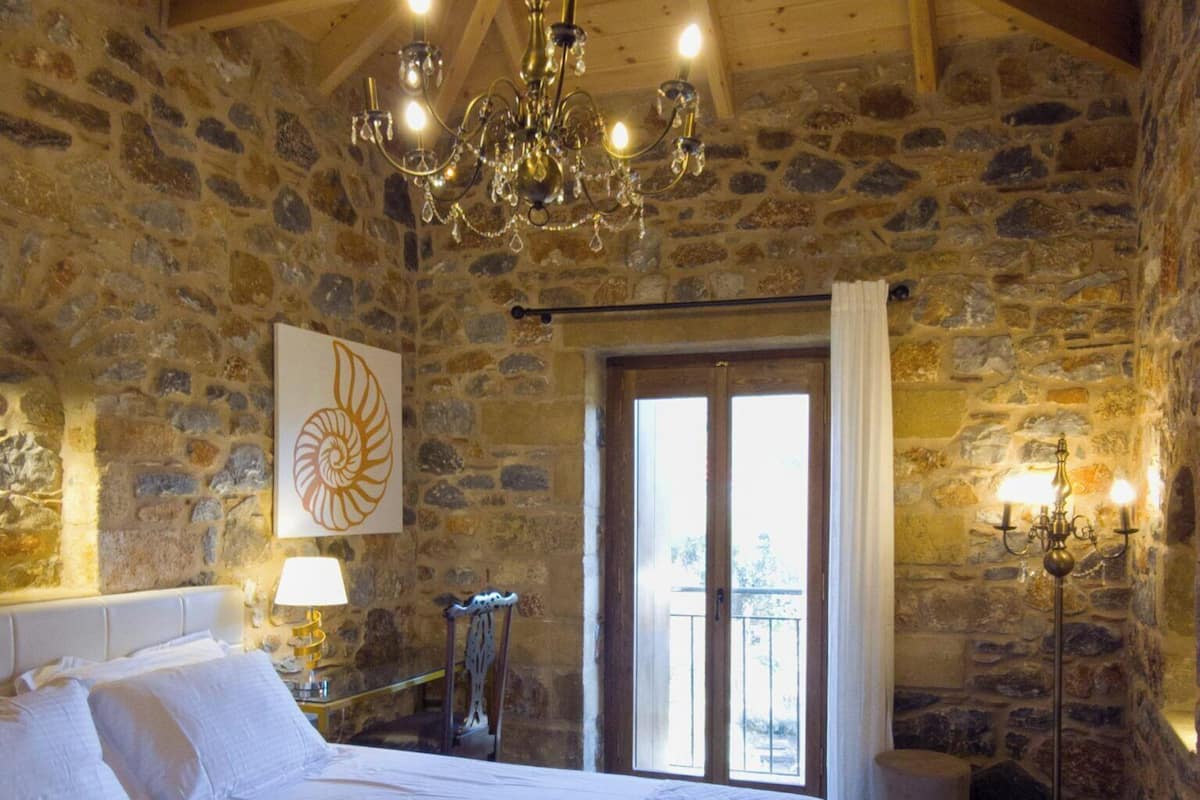 Une chambre avec un grand lit, des murs en pierre, un lustre et une vue sur l’extérieur, visible à travers une porte vitrée.