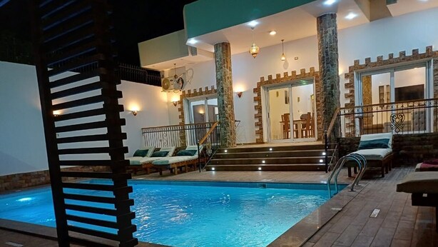 An indoor swimming pool with a wooden deck, a staircase leading to a balcony, and a stone pillar. - Twilight Palace (Luxor)