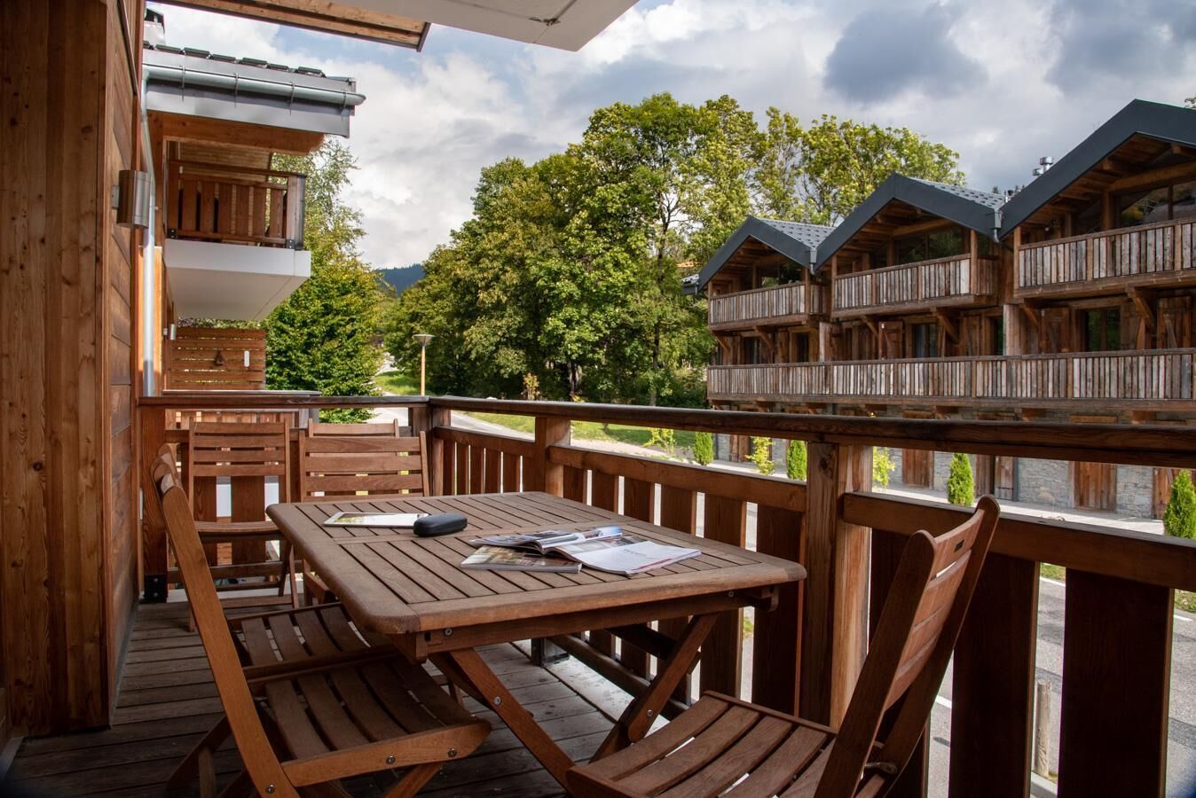 A wooden deck with a table and chairs, overlooking a forested area with multiple wooden buildings.