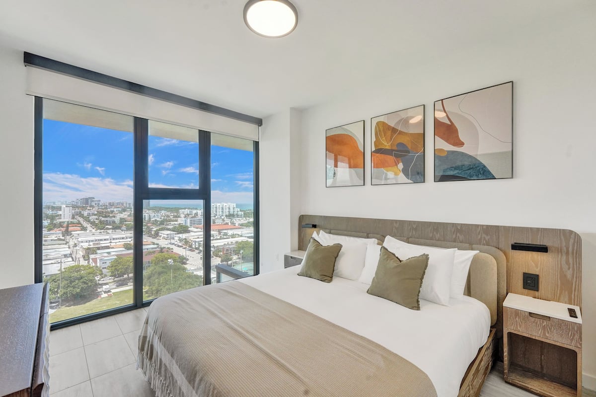 Ein modernes Hotelzimmer mit einem großen Bett, einem Schreibtisch und Blick auf die Stadt durch raumhohe Fenster.
