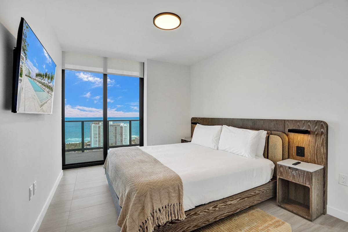 Ein Schlafzimmer mit einem großen Bett, einem Nachttisch, einem an der Wand befestigten Fernseher und einem Balkon mit Blick auf das Meer und die Gebäude.