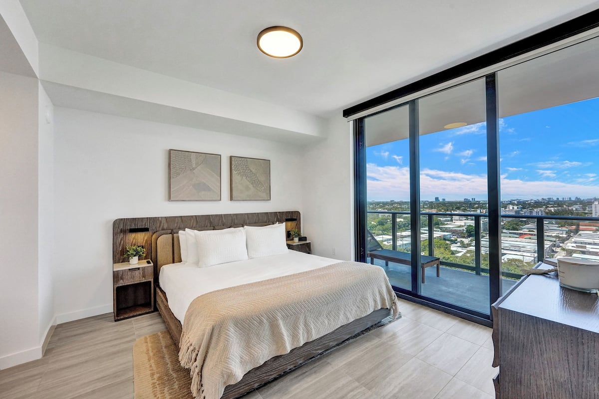 Ein modernes Schlafzimmer mit einem großen Bett, einem Nachttisch, einem Schreibtisch und Blick auf die Stadt durch große Fenster.
