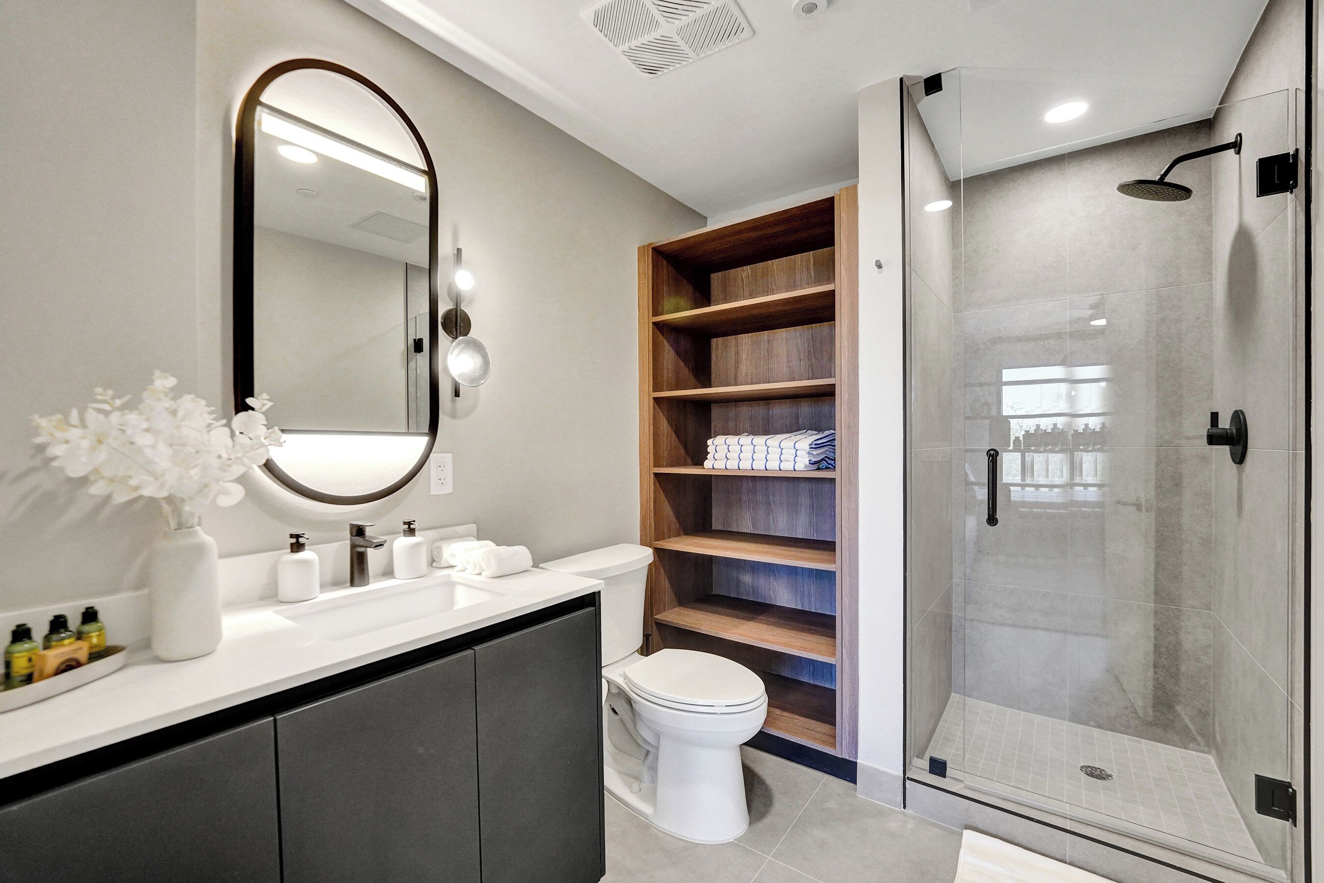 A modern bathroom with a white vanity, a large mirror, a glass shower enclosure, and a wooden shelving unit.