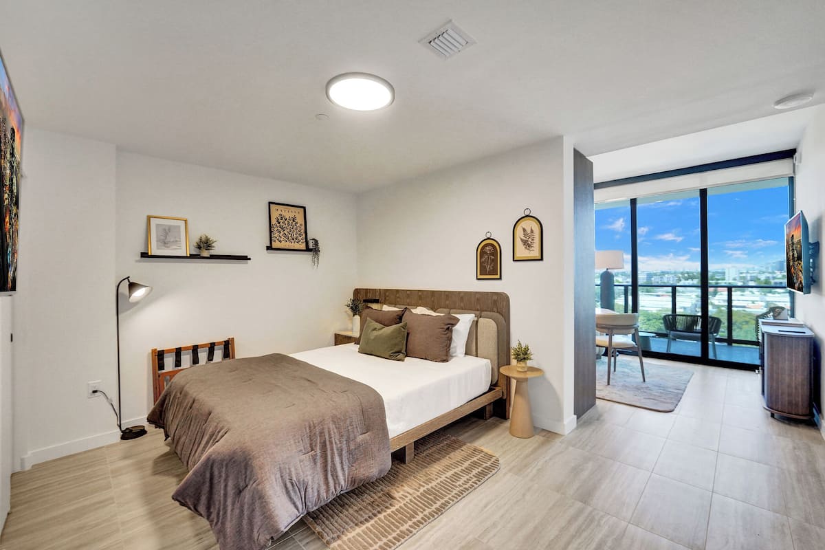 Ein modernes Schlafzimmer mit einem großen Bett, einem Nachttisch, einer Stehlampe und Blick auf die Stadt durch große Fenster.