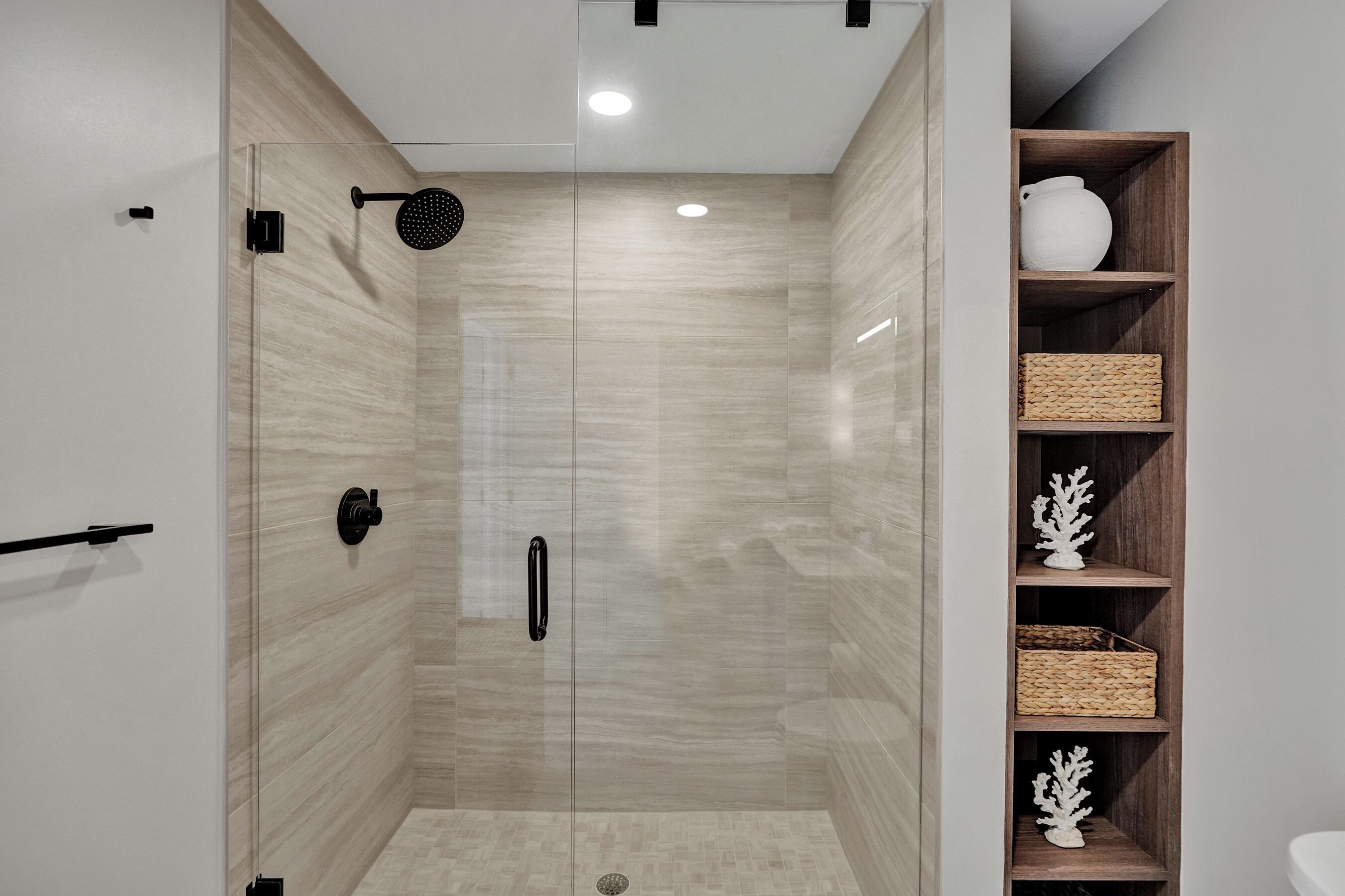 Une salle de bain moderne avec une douche à parois de verre, des étagères en bois ornées d’objets décoratifs et un lavabo blanc.
