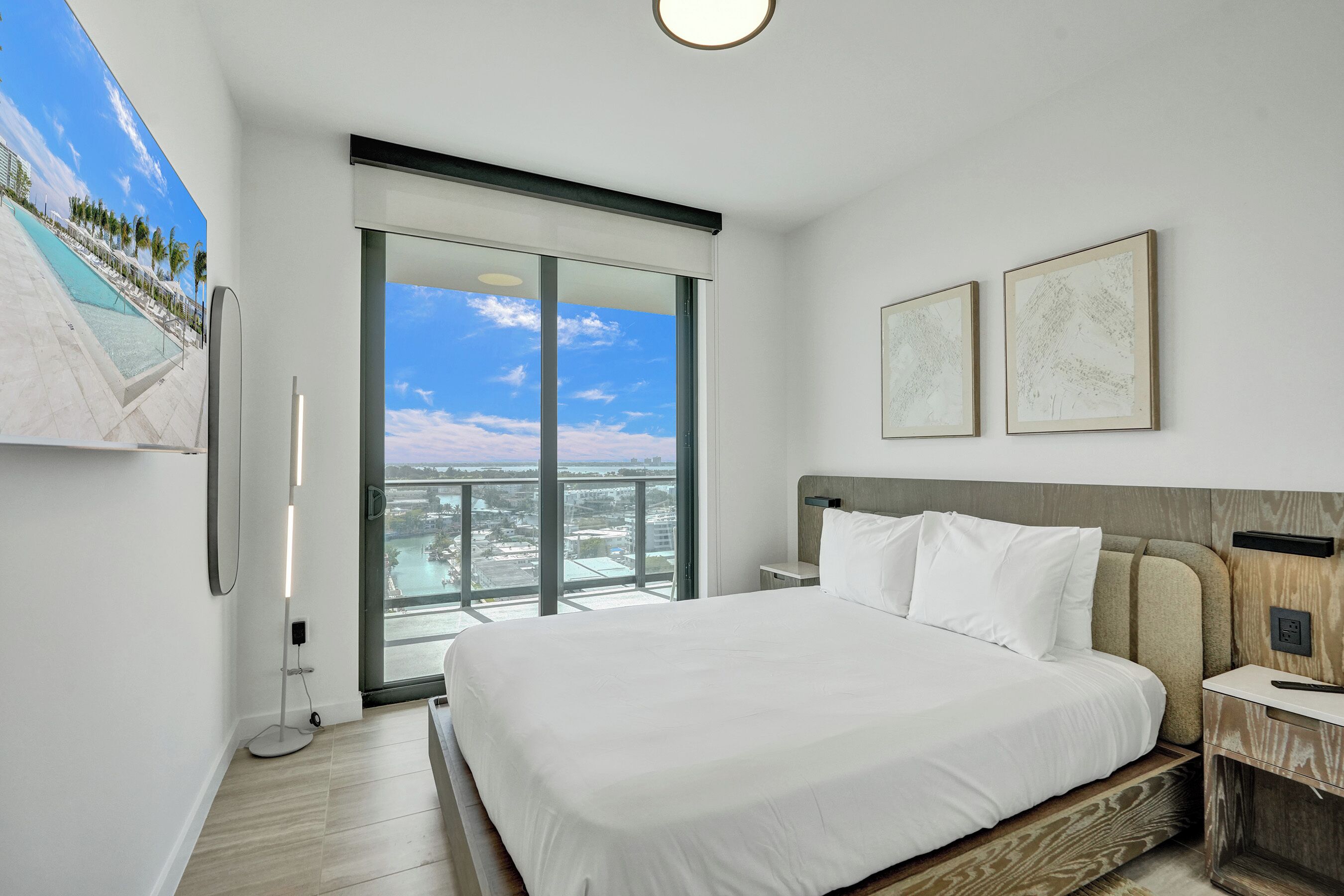 A modern hotel room with a large bed, a view of a cityscape through a sliding glass door, and two framed artworks on the wall.