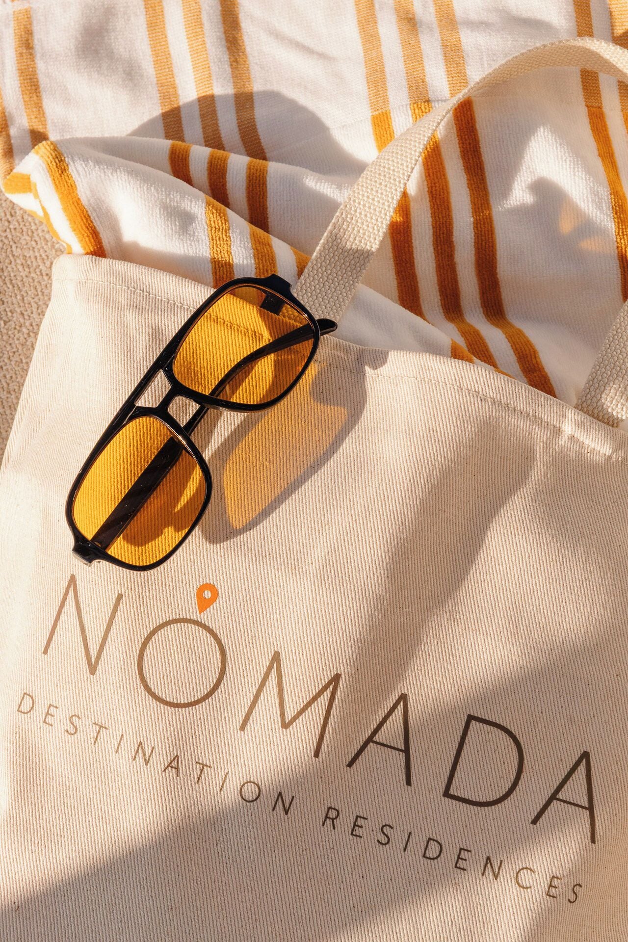 A white tote bag with 'NOMADA DESTINATION RESIDENCES' printed on it, featuring a pair of sunglasses.