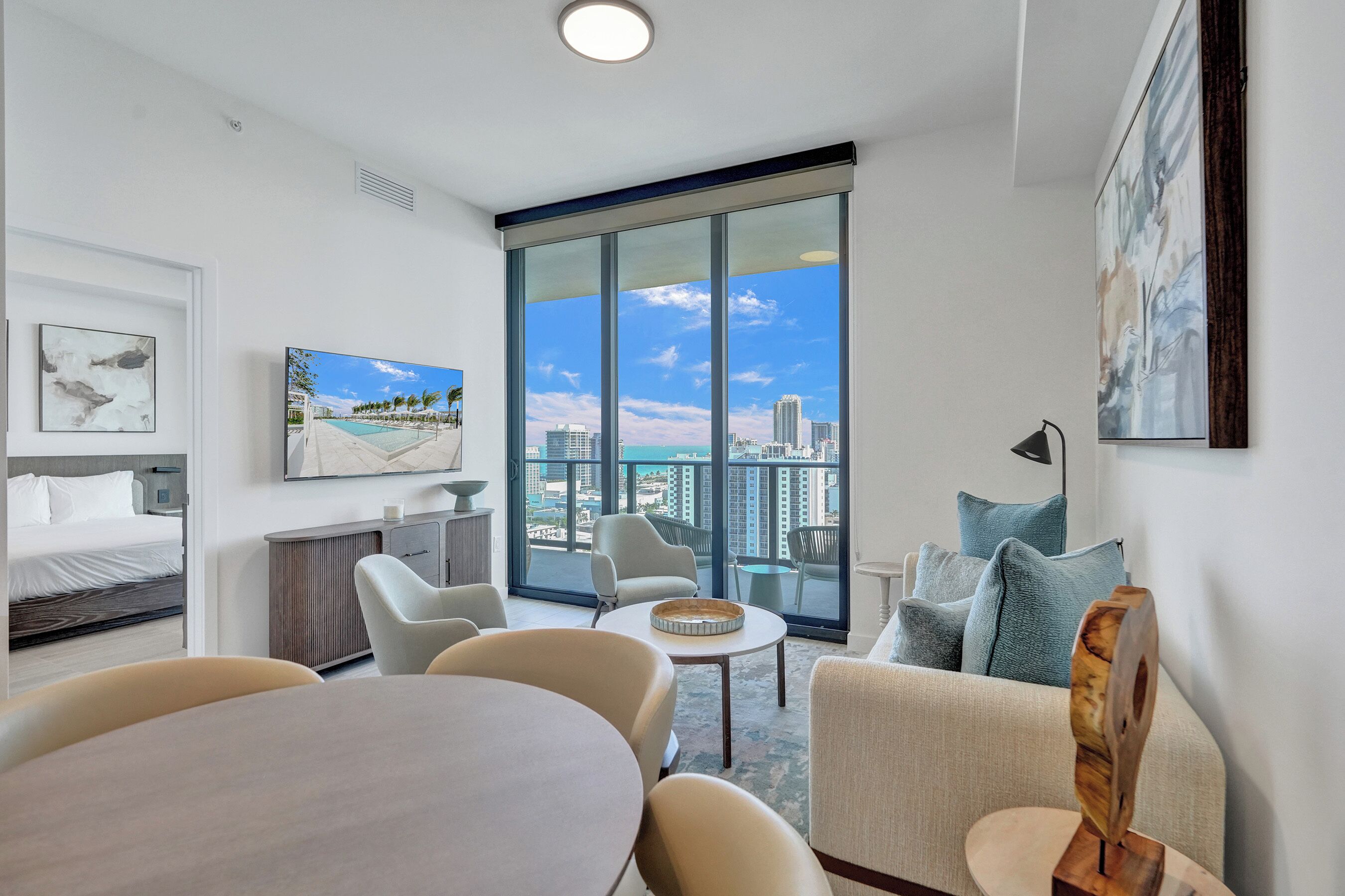 A modern hotel room with a large balcony, a bed, a dining area, and a seating area with a view of the city.