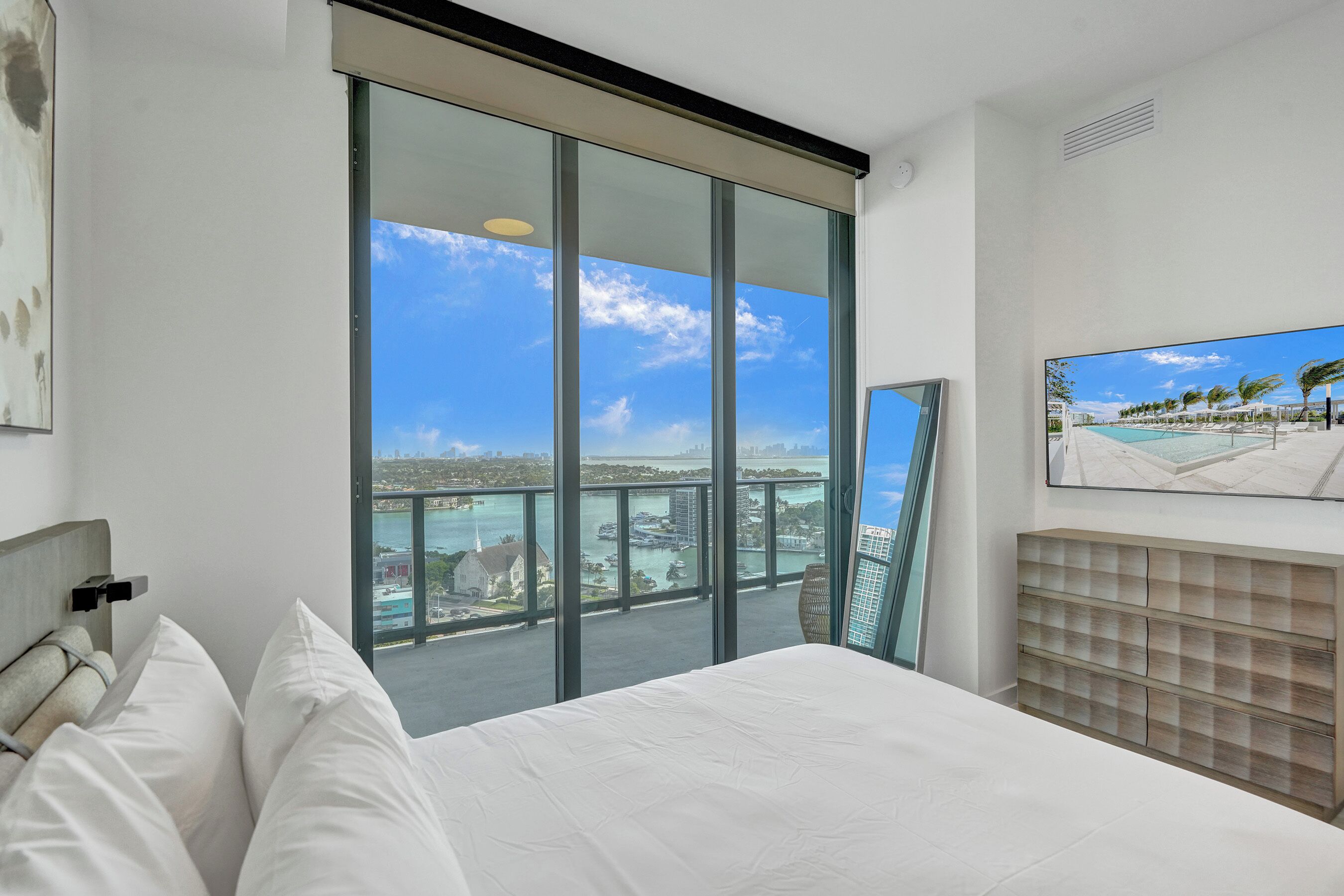 A hotel room with a large bed, a view of the city through floor-to-ceiling windows, and a mounted artwork on the wall.